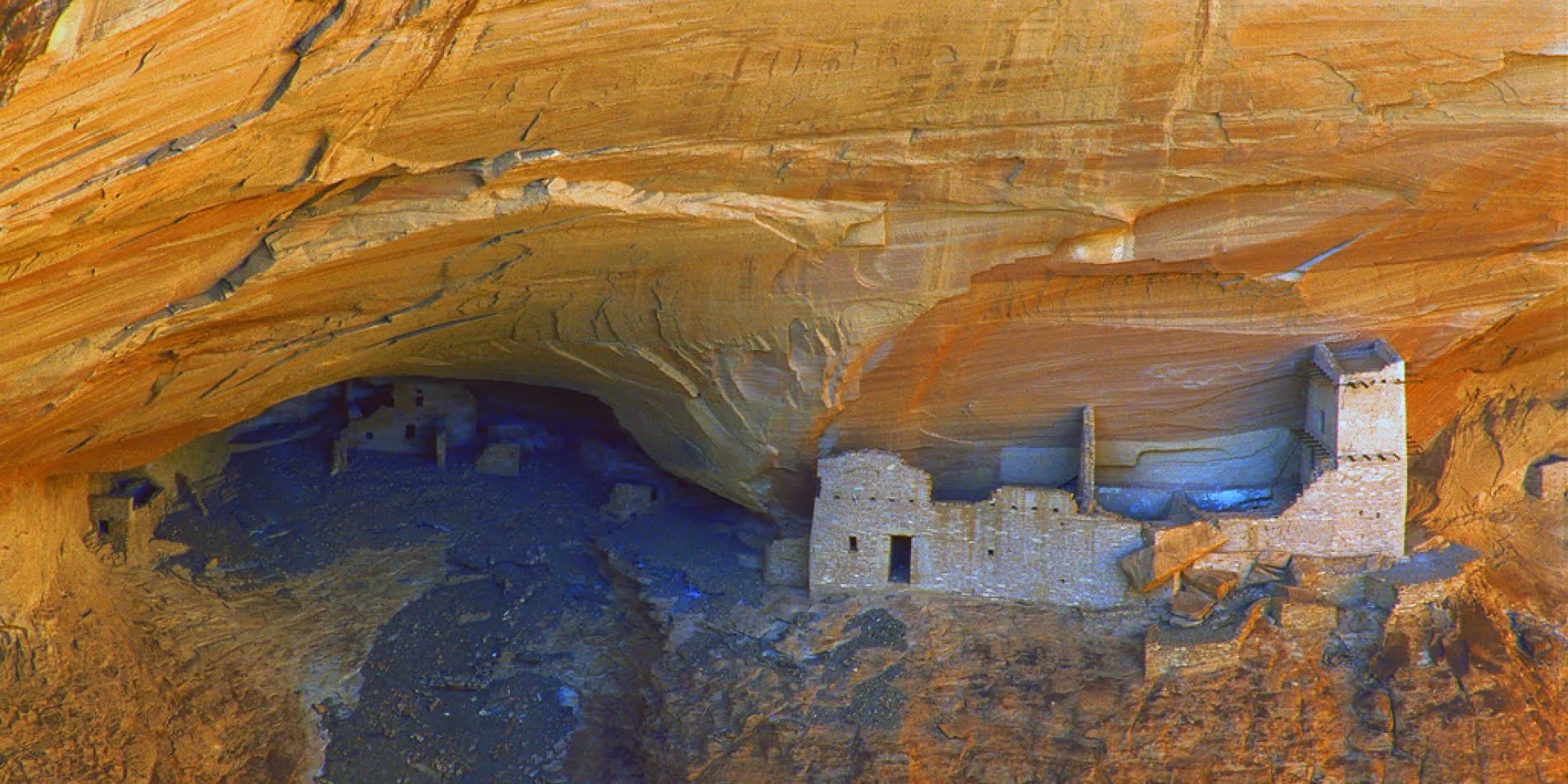 Ancient cliff dwellings built into a large sandstone cave wall with a tower and multiple stone structures.